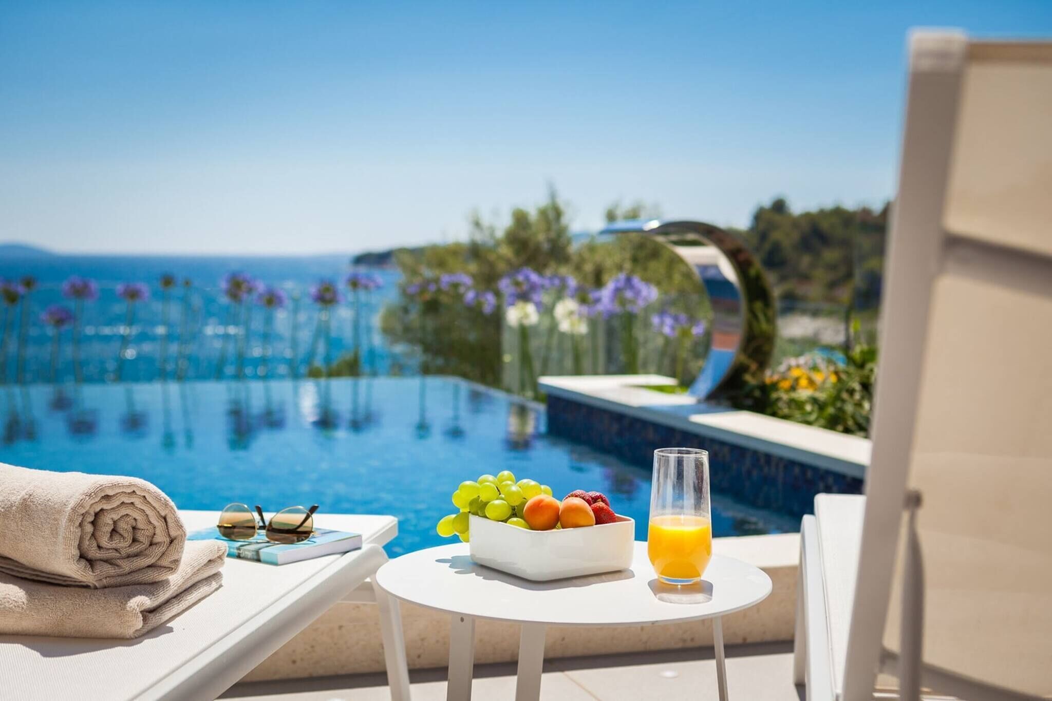 A poolside setting with a table, a bowl of fruit, a glass of orange juice, and a rolled towel. - Luxury Seafront Villa Blue Heaven Ciovo with Pool (Okrug Gornji)