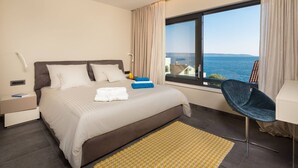 A hotel room with a bed, a chair, a nightstand, a window with a view of the sea, and a small table. - Luxury Seafront Villa Blue Heaven Ciovo with Pool (Okrug Gornji)