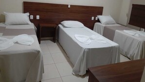Une chambre d’hôtel avec trois lits simples, chacun doté d’une tête de lit en bois et de draps blancs. Une table de chevet en bois se trouve entre les lits, et une petite table est placée dans un coin de la pièce.