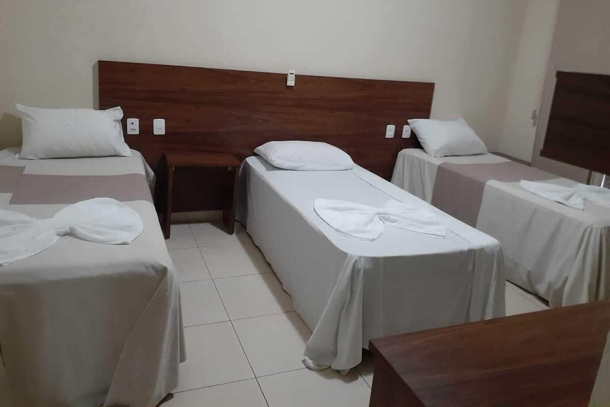 A hotel room with three single beds, each with a wooden headboard and white bedding. There is a wooden nightstand between the beds, and a small table in the corner.