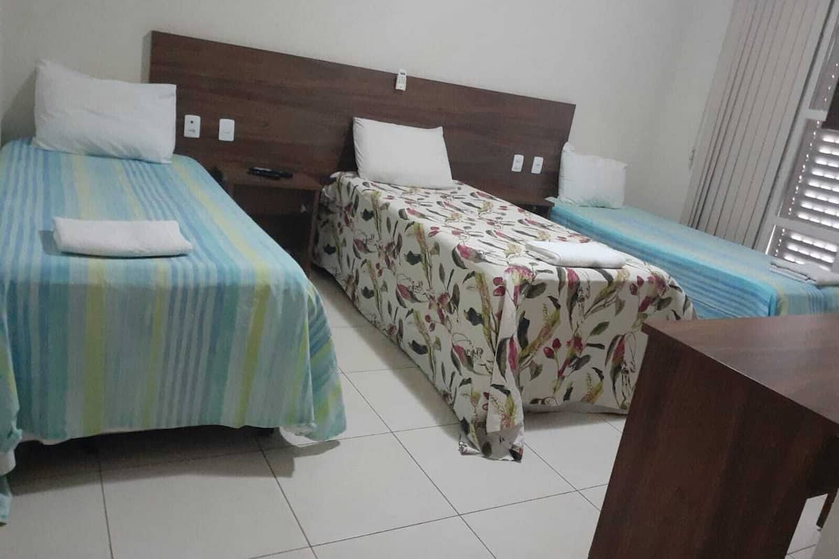 A hotel room with two single beds, each with a pillow and a blanket. There is a wooden headboard, a nightstand with a phone, and a window with blinds.