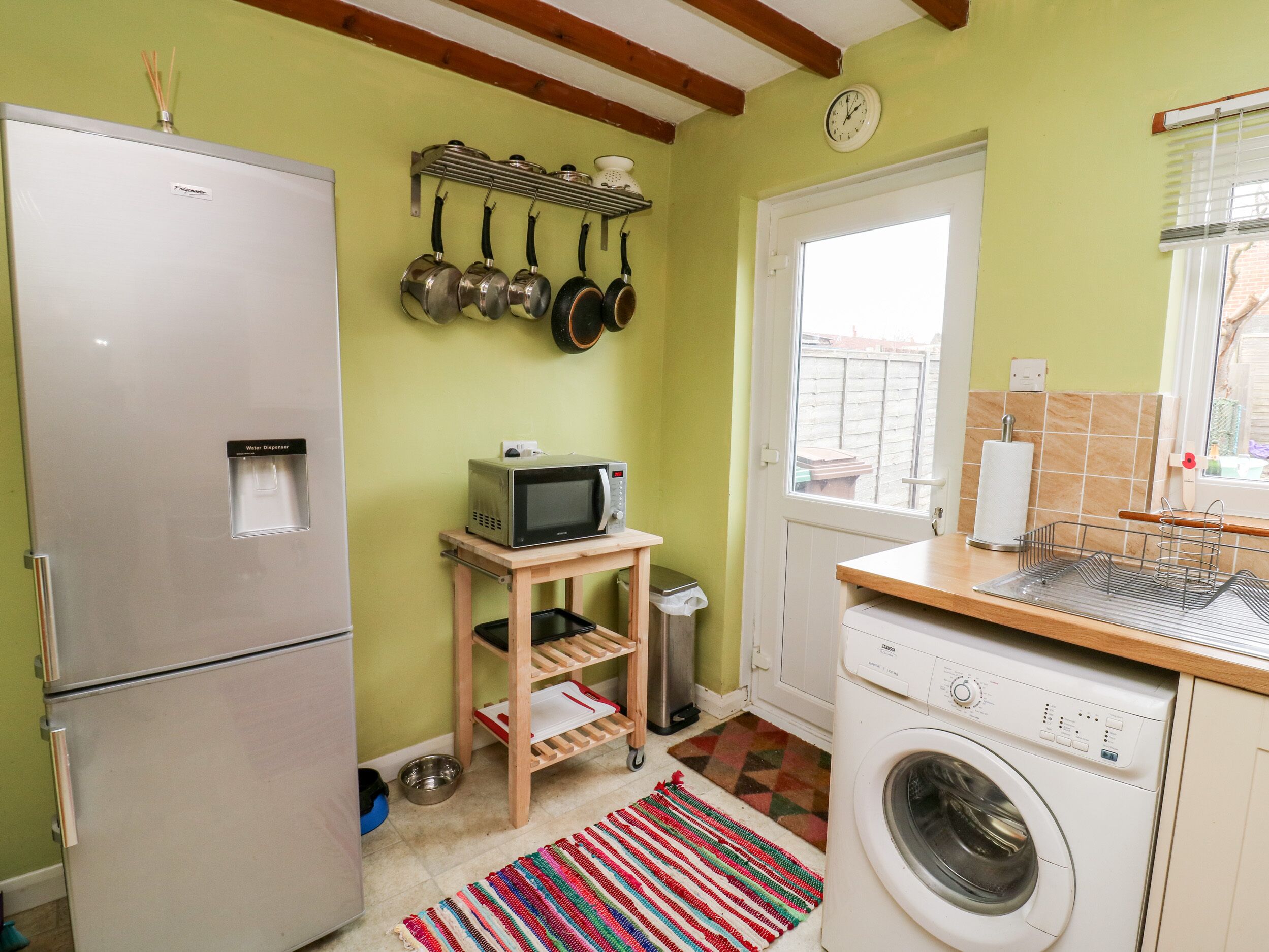 Une cuisine compacte équipée d’un réfrigérateur, d’un four à micro-ondes et d’un lave-linge. On y trouve une étagère en bois où sont suspendues des casseroles, ainsi qu’une petite table sur laquelle est posé un four à micro-ondes.