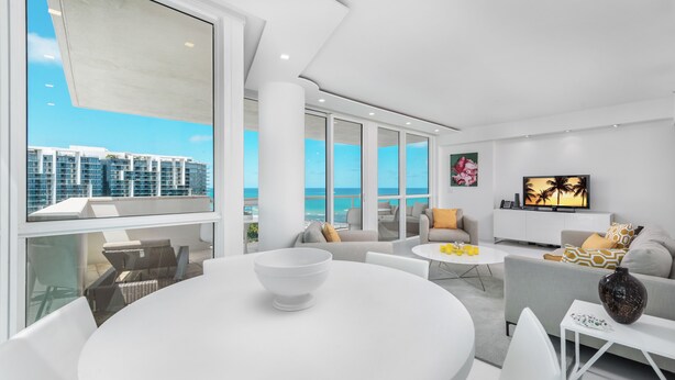 A modern living room with a large window, a round dining table, and a sofa with cushions. - Oceanview Private Condo at The Setai -190102 (Miami Beach)
