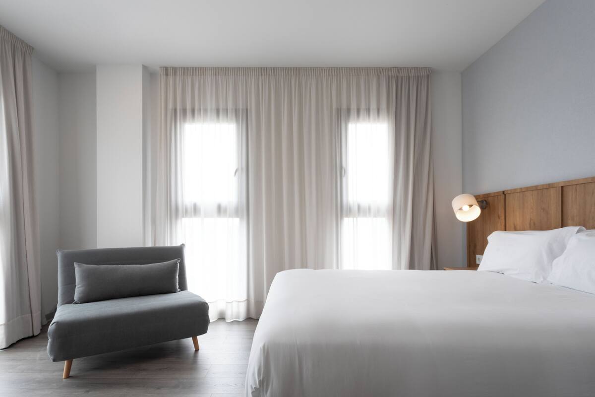 A hotel room with a bed, a chair, a nightstand with a lamp, and large windows with curtains.