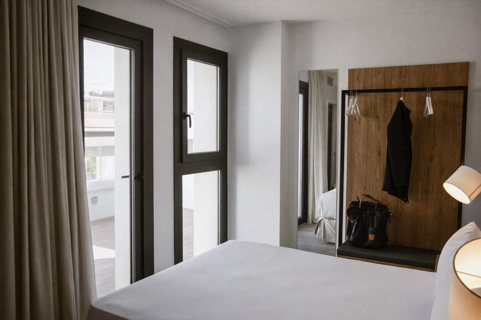 Hotelkamer met een bed, een houten garderobe, een spiegel, een kapstok en een raam met gordijnen.