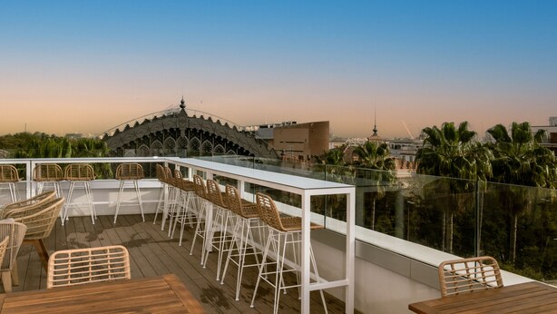 A rooftop bar with white bar stools and a wooden table, overlooking a cityscape and palm trees. - Petit Palace Vargas (Seville)