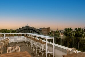 A rooftop bar with white bar stools and a wooden table, overlooking a cityscape and palm trees. - Petit Palace Vargas (Seville)