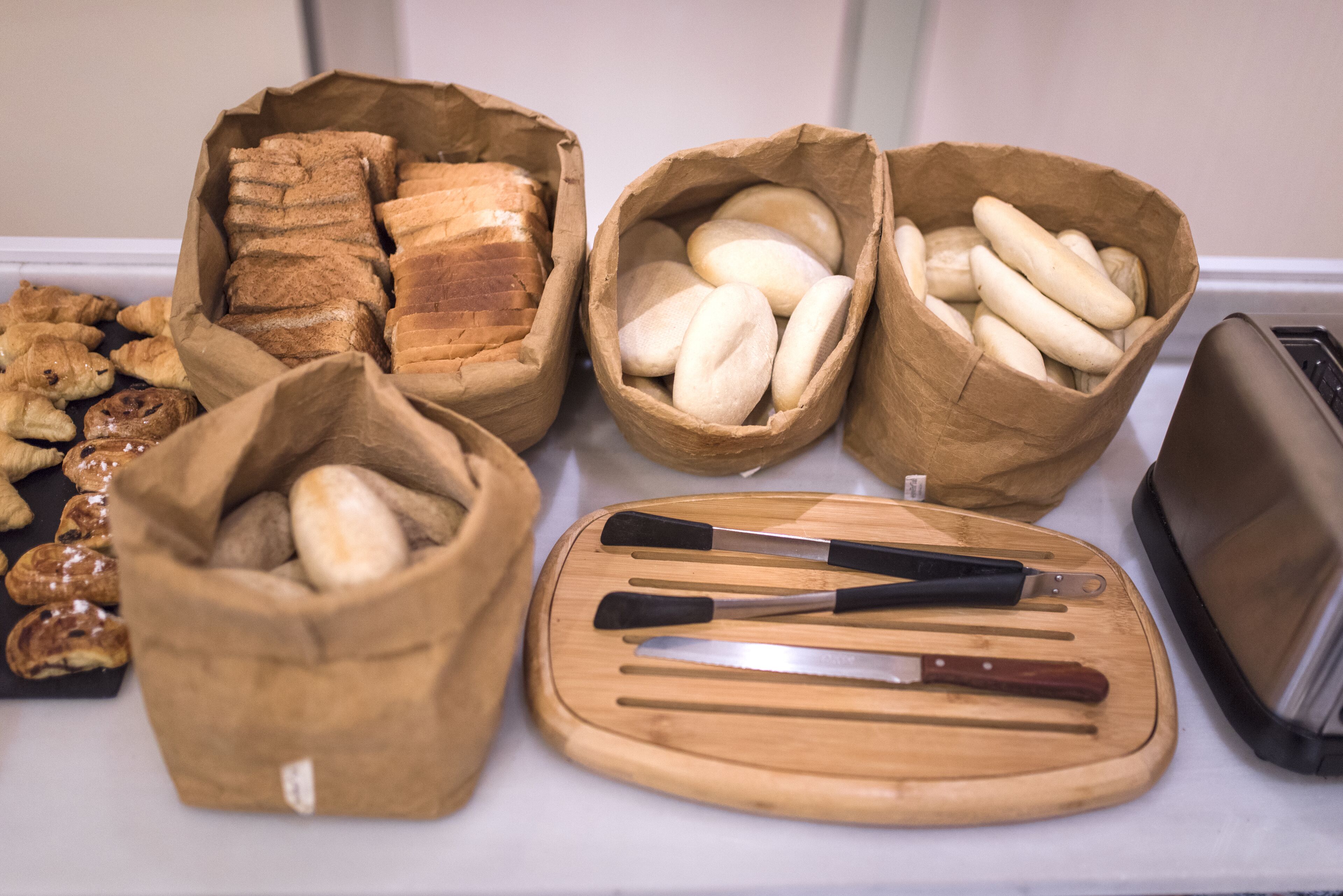 A display of various breads and pastries in paper bags, with a cutting board and knives in the foreground.
