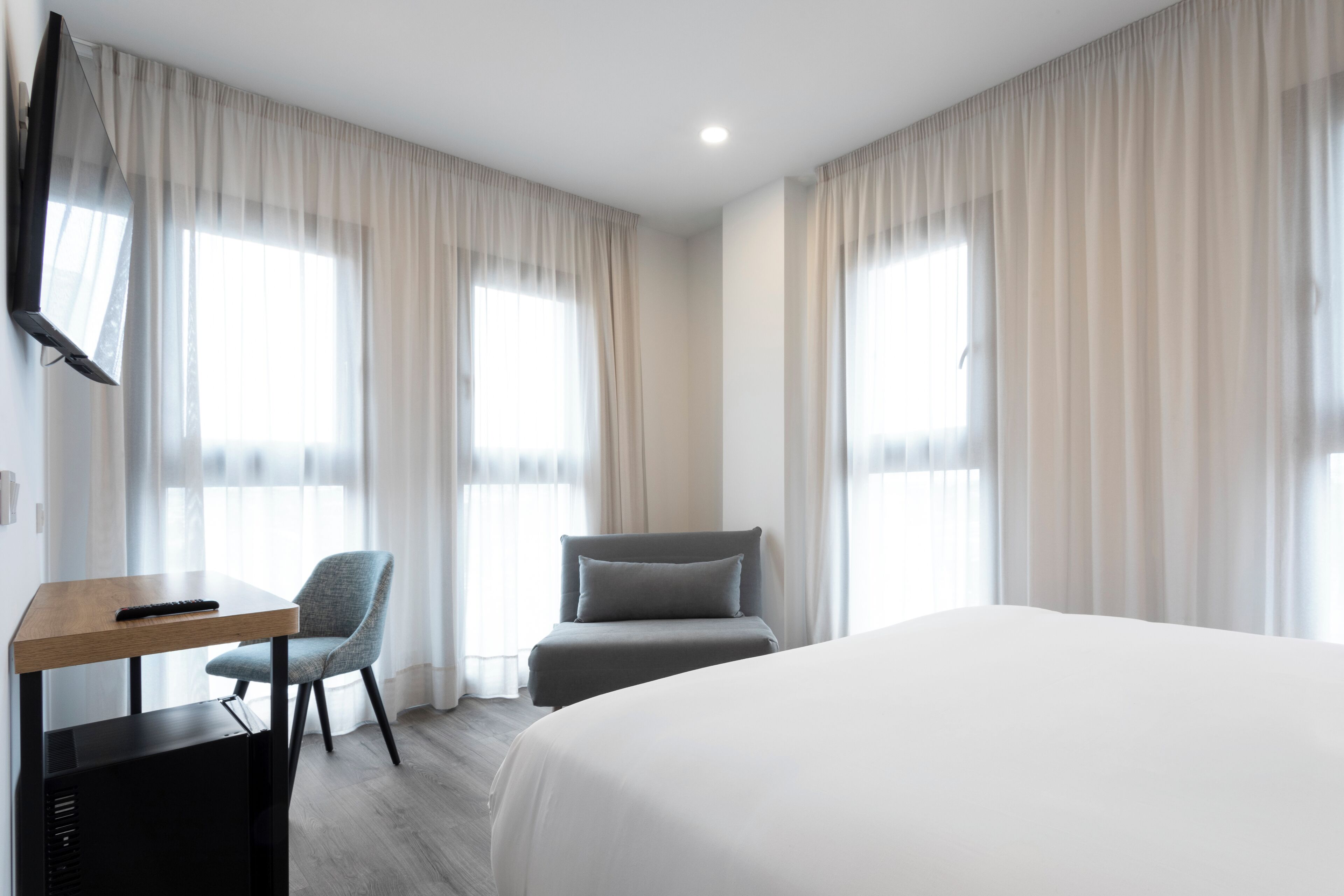 A hotel room with a bed, a desk with a chair, a TV, and large windows with curtains.