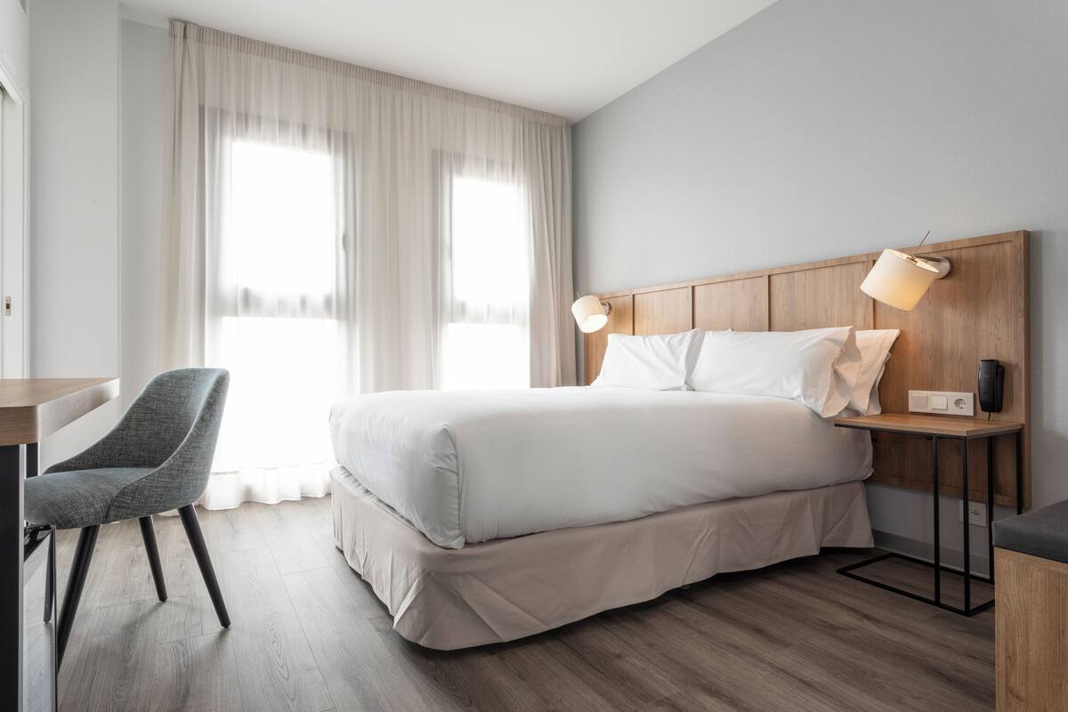 A hotel room with a bed, a desk, a chair, a bedside table with a lamp, and a window with curtains.