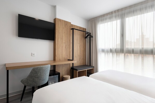 Una habitación de hotel con una cama, un escritorio, una silla, un televisor montado en la pared y una ventana con cortinas translúcidas.