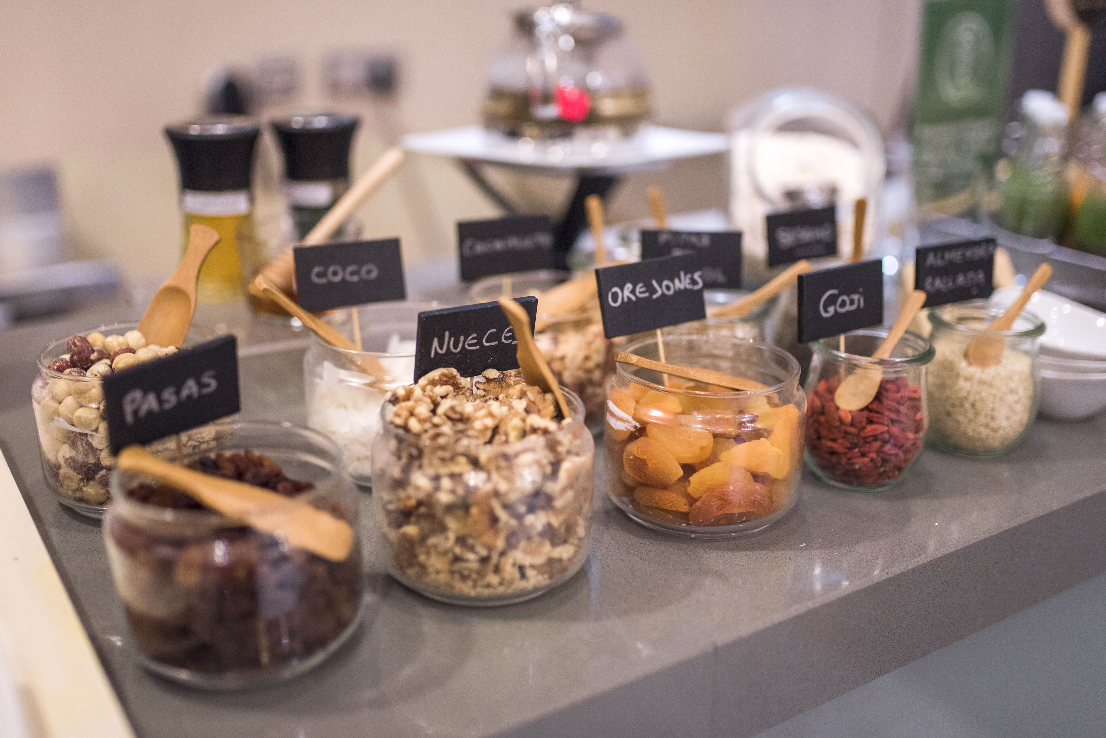 Diversos potes etiquetados com diferentes ingredientes como cacau, nozes e frutas secas, provavelmente em um café ou padaria.