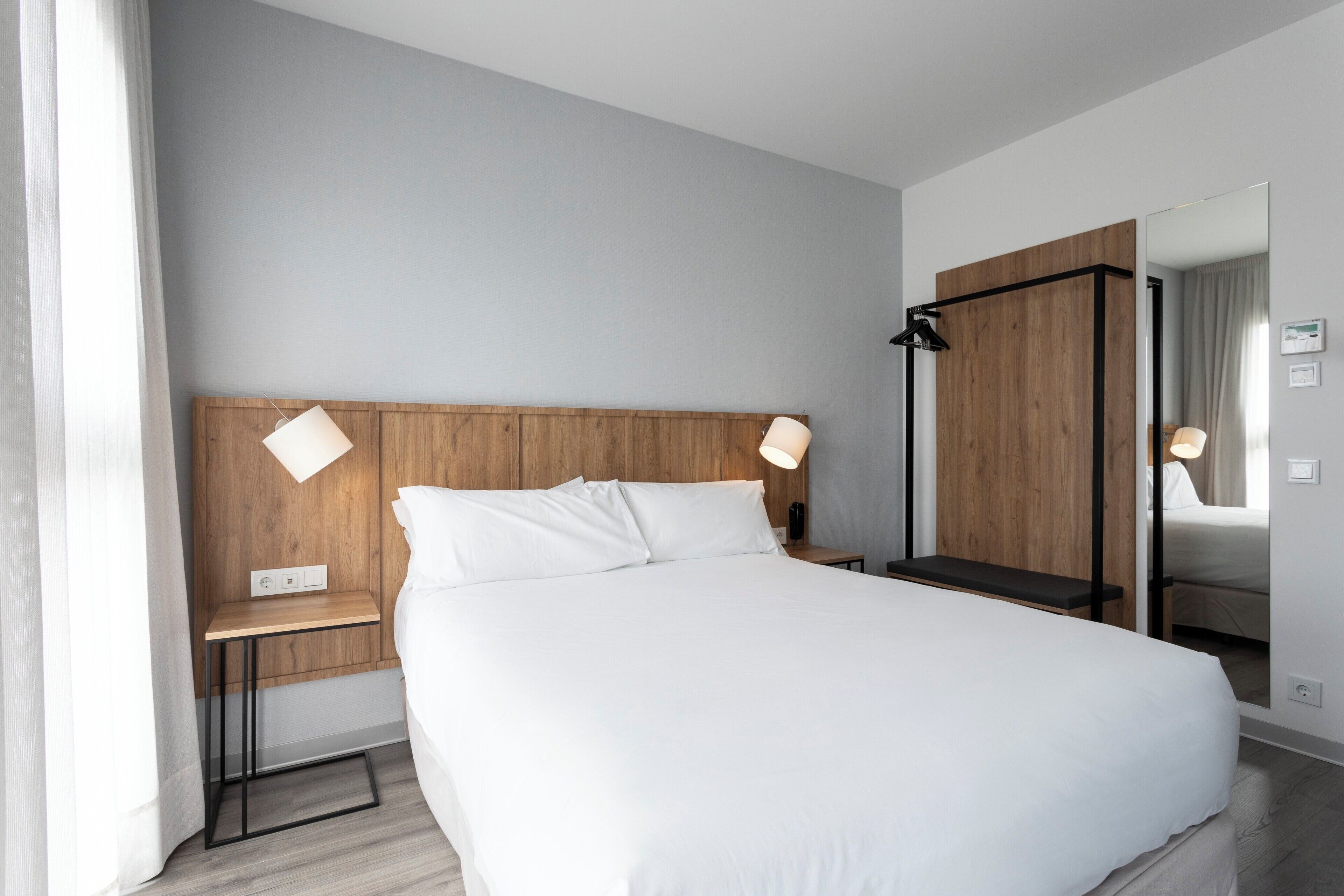 Una habitación de hotel moderna con una cama grande, mesitas de noche, un espejo y un cabecero de madera.