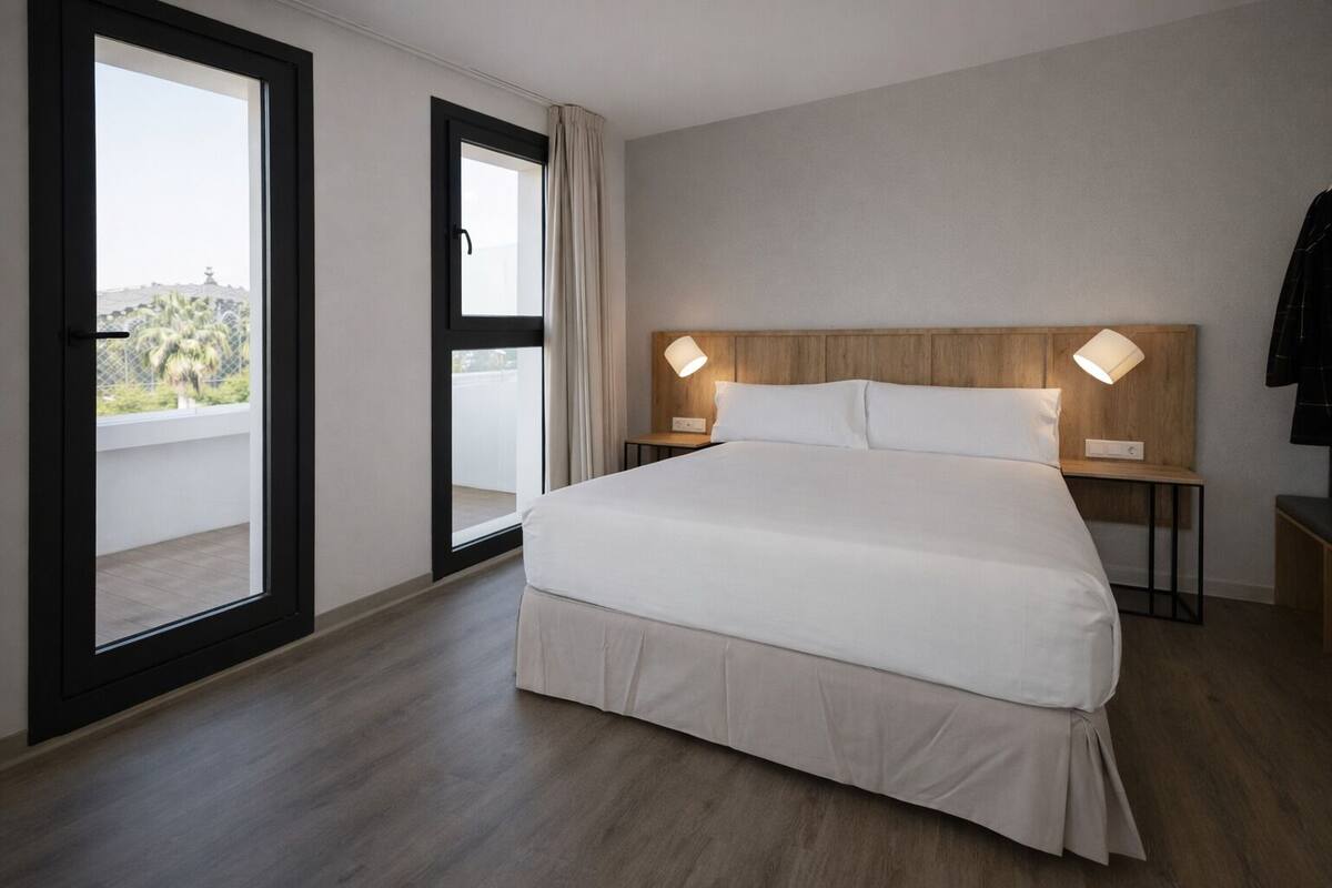 A hotel room with a large bed, a wooden headboard, two bedside lamps, a nightstand, a sliding glass door to a balcony, and a view of palm trees.