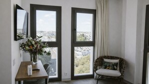 A room with two large windows, a wooden table with a vase of flowers, a wicker chair, and a view of a cityscape. - Petit Palace Vargas (Seville)
