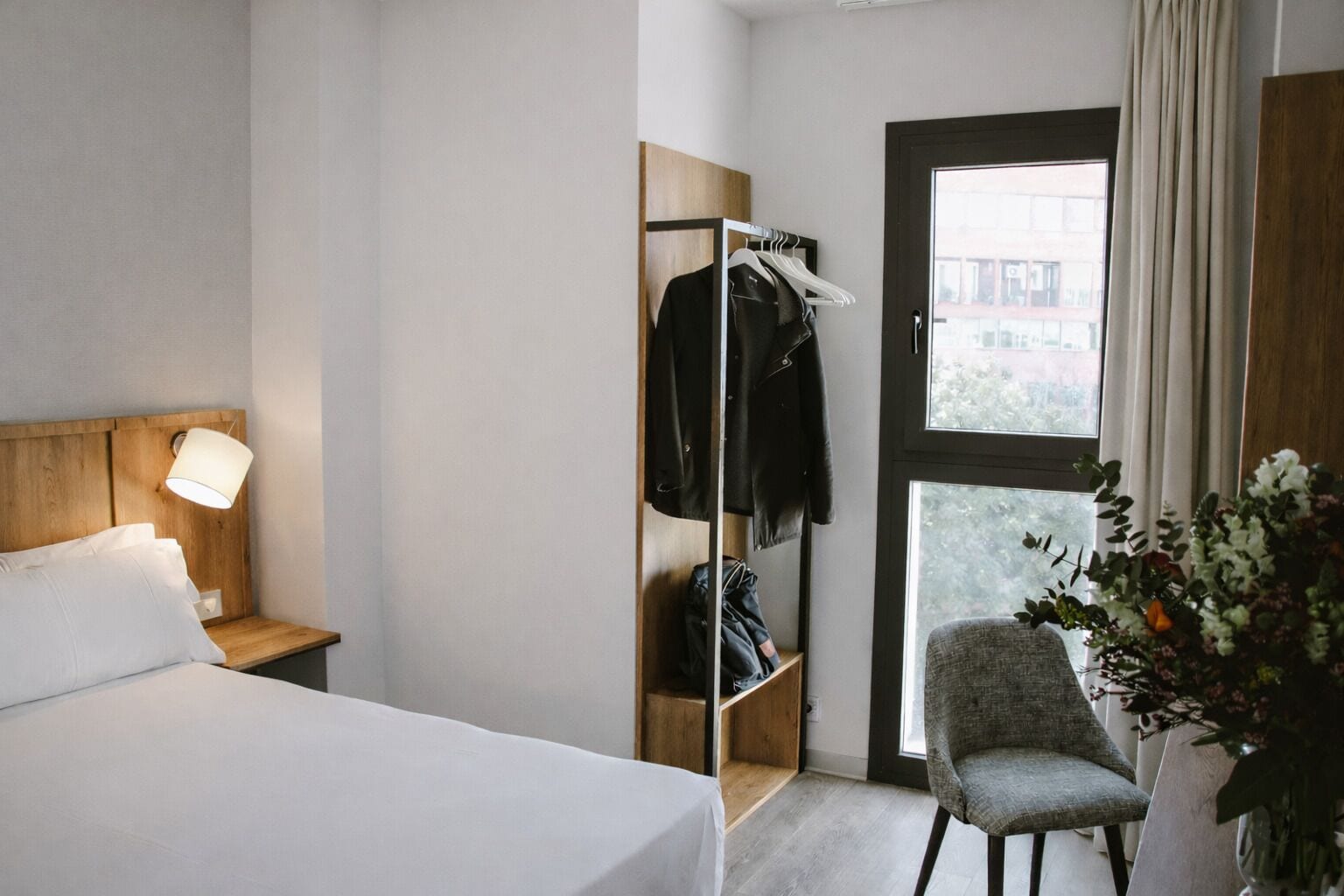 A hotel room with a bed, a nightstand with a lamp, a wardrobe, a chair, and a window with a view.