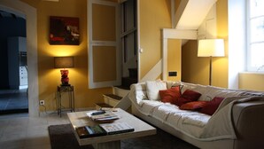 Een woonkamer met een witte bank, rode kussens, een bijzettafeltje met boeken, een lamp en een schilderij aan de muur.