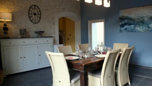 Een eethoek met een houten tafel voor zes personen, omringd door rieten stoelen. Links bevindt zich een witte kast met lades, en aan de muur hangt een klok.
