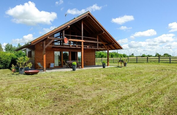 Una cabaña de madera con techo inclinado, ventanas grandes y un patio con mobiliario exterior.
