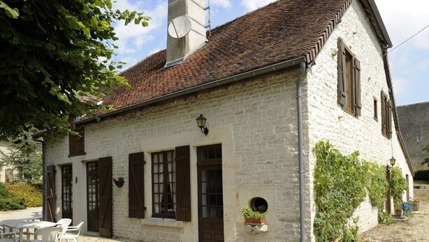 A stone building with a tiled roof, white walls, and brown shutters. There is a small round window on the side and a door with a window above it.