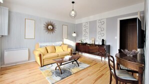 A living room with a yellow sofa, a wooden coffee table, a dining table with chairs, a TV, and a decorative mirror on the wall.
