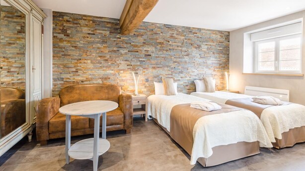 Une chambre d’hôtel avec un mur en pierre, deux lits, un canapé, une petite table et une grande fenêtre.