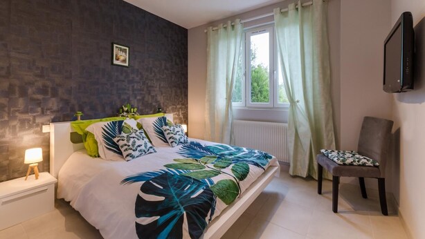 Un dormitorio con cama, mesita de noche, una silla, ventana con cortinas y televisor montado en la pared.
