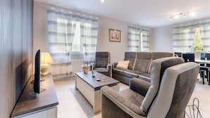 Una sala de estar moderna con un sofá, sillones y una mesa de centro. Hay un televisor sobre un mueble bajo, una lámpara y un cuadro en la pared.
