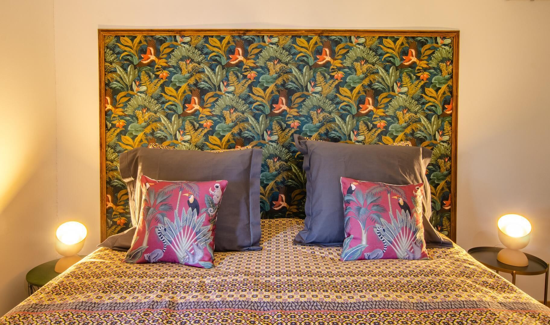 Uma cama com colcha estampada, duas almofadas com motivos tropicais e uma cabeceira com padrão floral detalhado.