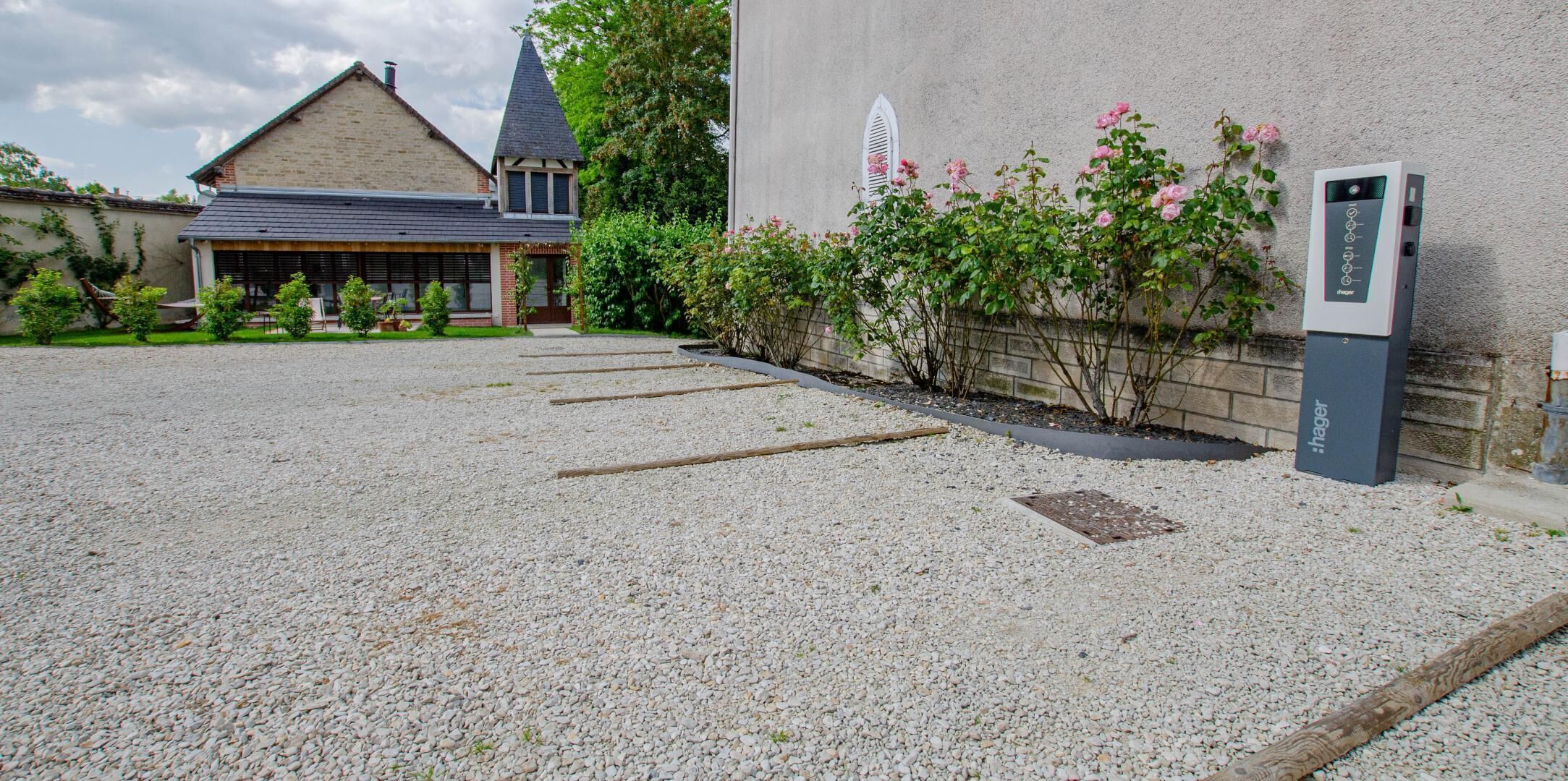 Une allée de gravier mène à un bâtiment de style architectural traditionnel, agrémenté d’un petit jardin où poussent des rosiers roses, et doté d’une borne de recharge moderne pour véhicules électriques.