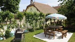 A patio area with wooden picnic tables and a covered seating area, surrounded by lush greenery and a building with a tiled roof.