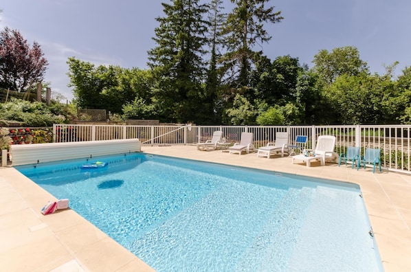 A rectangular swimming pool with clear blue water, surrounded by a white fence. There are several lounge chairs and a small table with a blue umbrella.