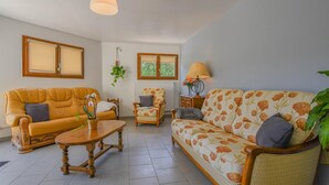Ein Wohnzimmer mit einer Couch, einem Sessel und einem Tisch. Es gibt Fenster mit Jalousien, eine hängende Pflanze und eine Lampe auf einem Beistelltisch.