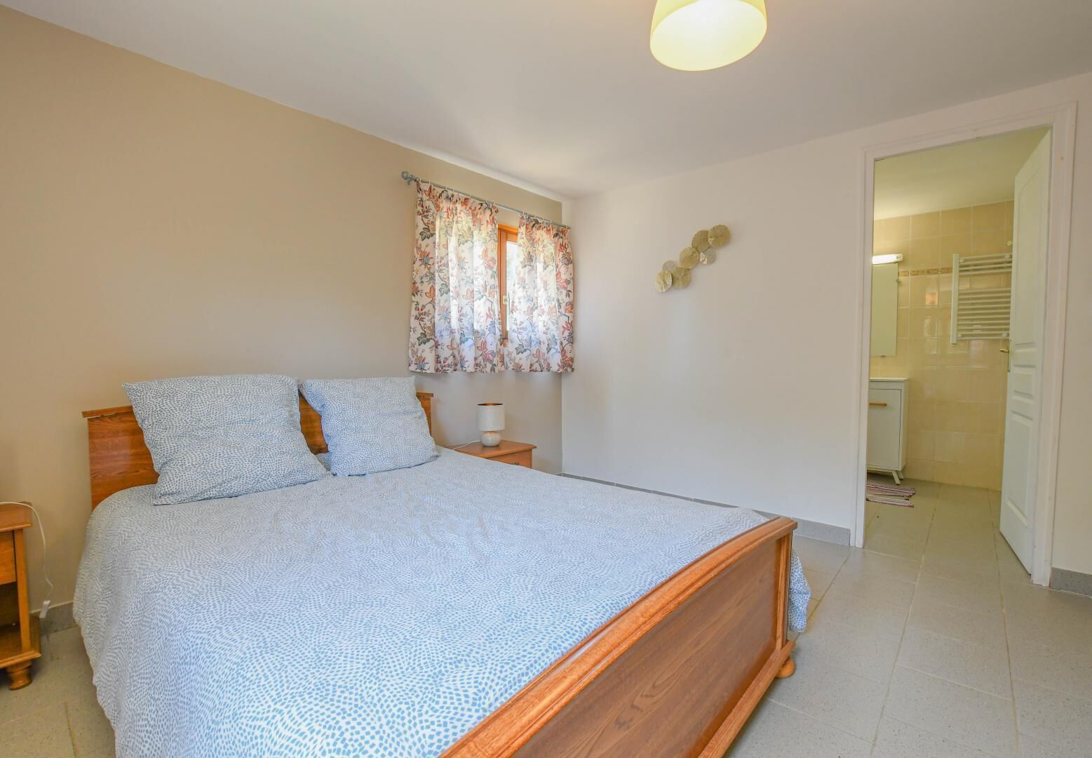 A bedroom with a wooden bed, a nightstand, a lamp, a floral curtain, and a bathroom visible through an open door.