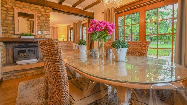 A dining area with a glass table, wicker chairs, a fireplace, and a view of greenery outside.