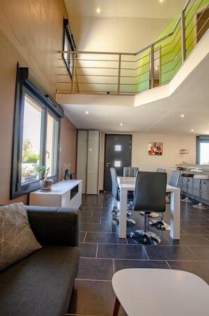 Een moderne woonkamer met een bank, een eettafel en stoelen. Er is een trap die naar een hoger gelegen verdieping leidt.