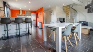 A modern kitchen with a white countertop and orange walls, a dining area with a white table and grey chairs, and a staircase with wooden railings. - Chez Mimi - Plessis Barbuise (PLESSIS BARBUISE)