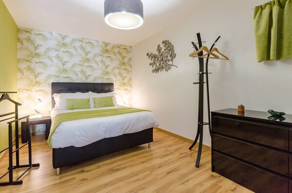 A bedroom with a bed, bedside table, lamp, wall art, coat rack, and a dresser.