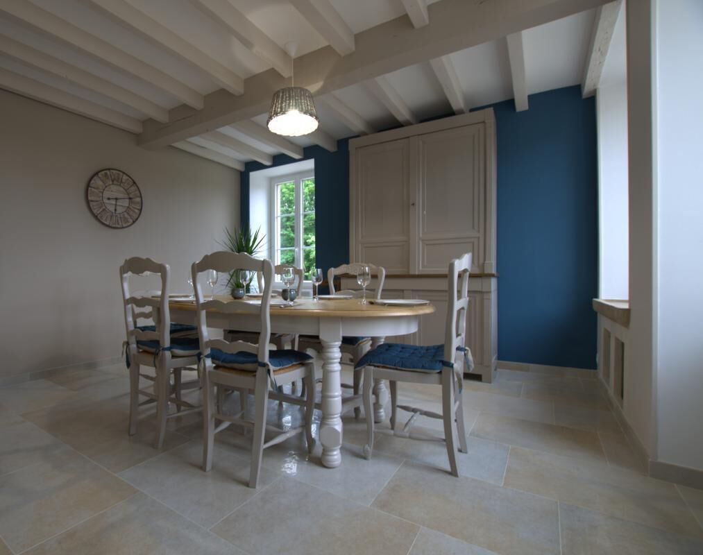 A dining area with a round table and six chairs, a chandelier, a clock on the wall, and a view of the outdoors through a window.