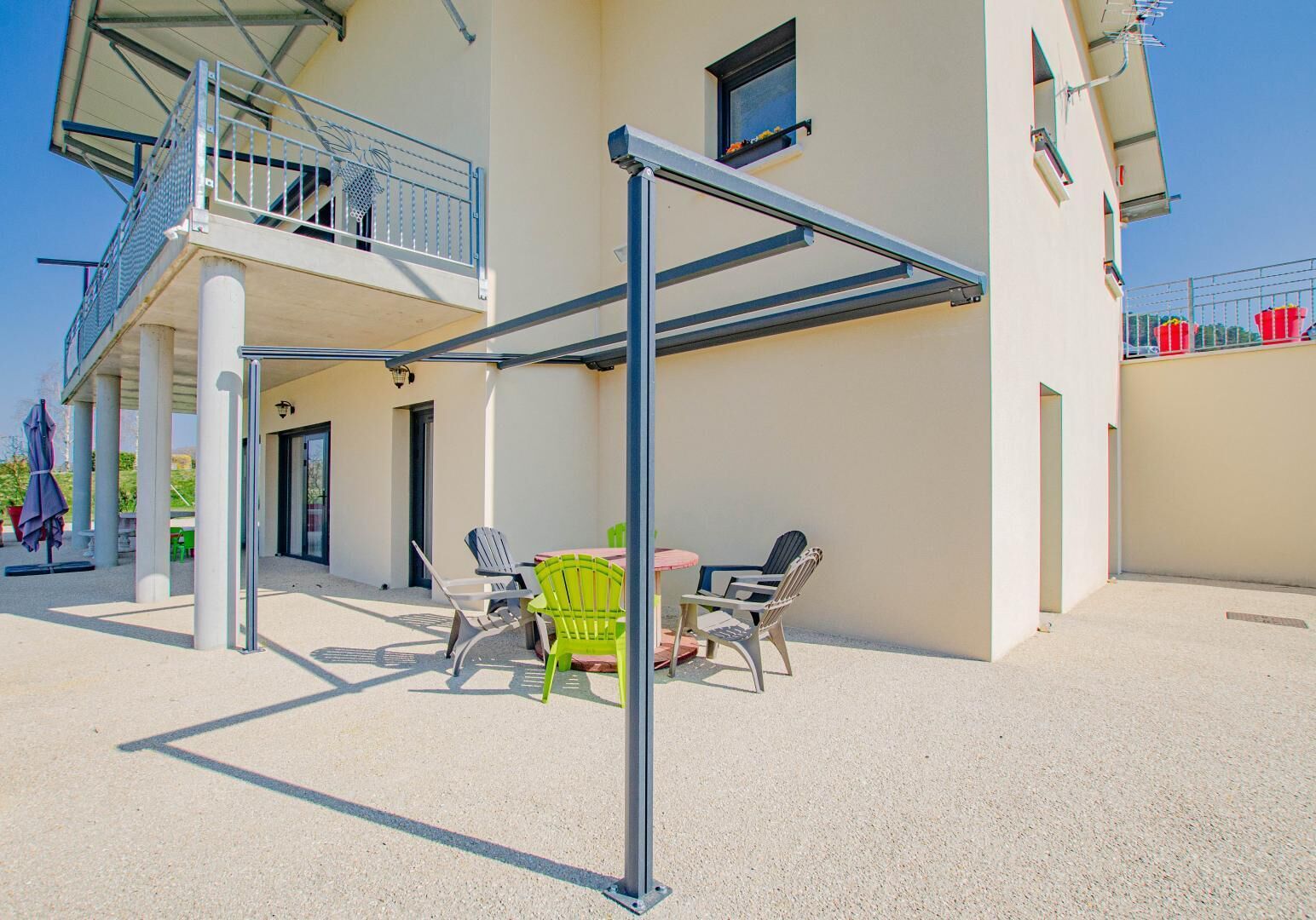 Un espace extérieur abrité, doté d’une table et de chaises, d’une structure métallique et d’un bâtiment avec un balcon.