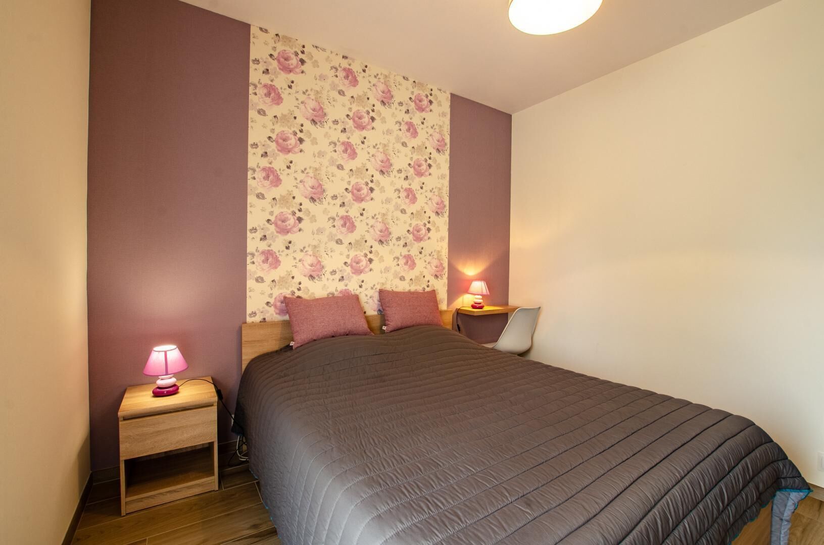 A bedroom with a bed, two bedside tables with lamps, a floral wallpaper, and a wooden floor.