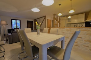 Un espace repas moderne avec une table et des chaises aux couleurs claires, un vase avec des fleurs et, au fond, une cuisine.