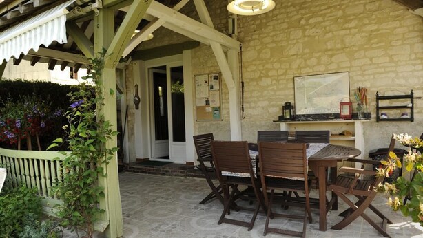 A covered patio with a stone wall, wooden furniture, and a small table with chairs. - La Salle de Danse - Eaux Puiseaux (EAUX PUISEAUX)