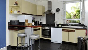 A compact kitchen with a stove, oven, and dishwasher, a window with a view of greenery, and a clock on the wall. - La Salle de Danse - Eaux Puiseaux (EAUX PUISEAUX)