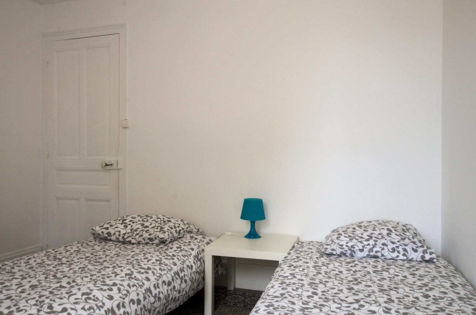 A small, simple room with two single beds, a small table with a lamp, and a white door.