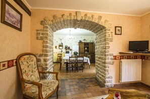 A living room with a stone archway leading to a kitchen. There is a wooden chair with floral upholstery and a table with a wine glass.