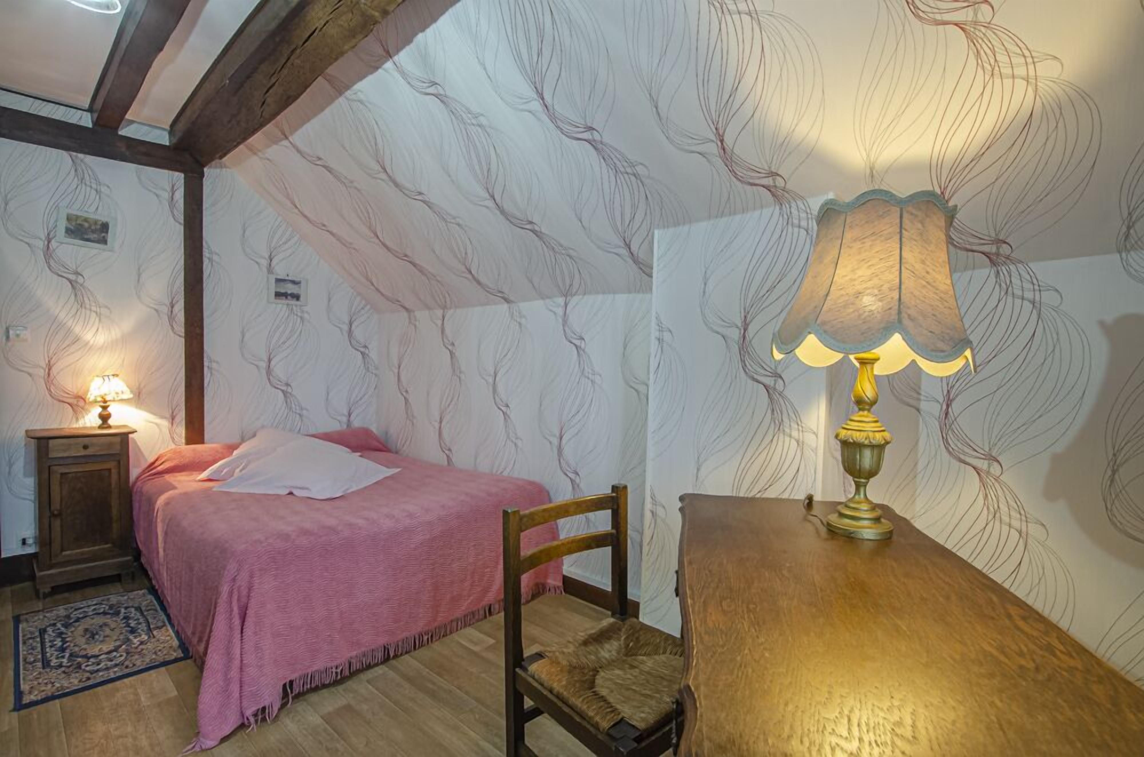 A bedroom with a bed, a chair, a table, a lamp, and a patterned wallpaper.