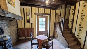 A room with a fireplace, a round table with chairs, a wooden bench, a staircase, and a door with a window. - House (BLIGNY)