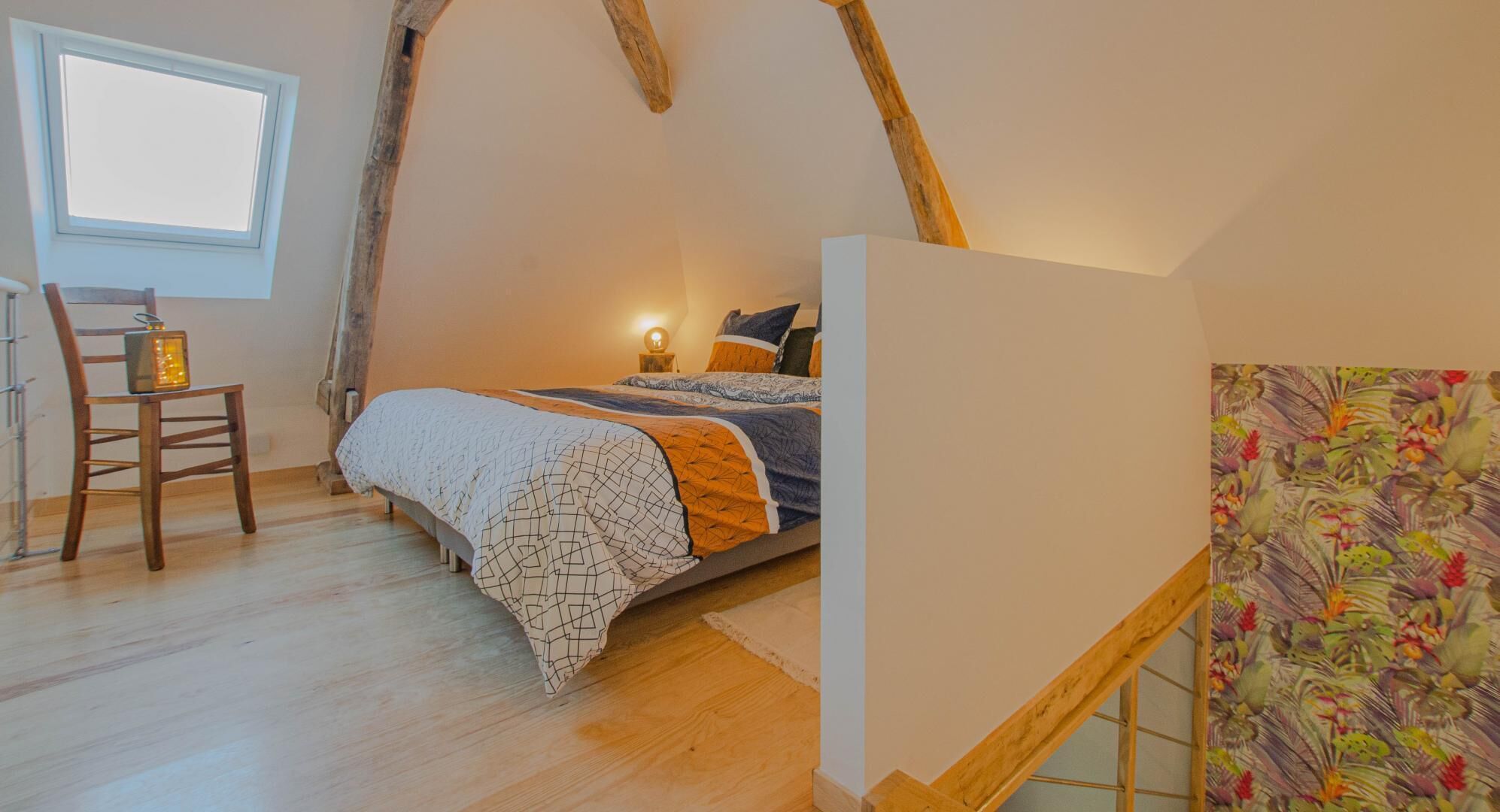 Ein Schlafzimmer mit schrägen Wänden, einem Bett mit gemusterten Textilien, einem Sessel mit Laterne und einem Fenster.