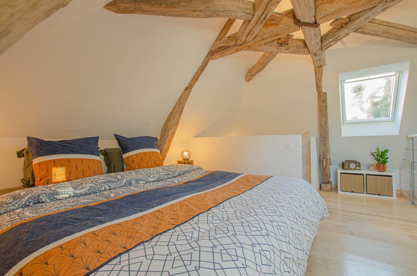 Ein Schlafzimmer mit schrägen Wänden, einem großen Bett mit gemusterten Betttextilien, einem Nachttisch mit Lampe und einem Fenster mit Blick auf Bäume.